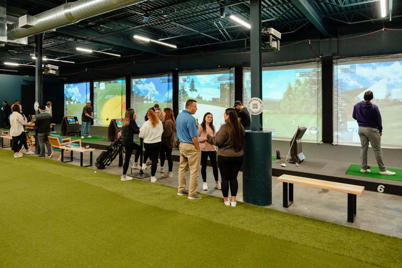 Golfplay People gather inside an Indoor Golf facility featuring several golf simulators, artificial turf flooring, and screens displaying virtual golf courses. Indoor Golf