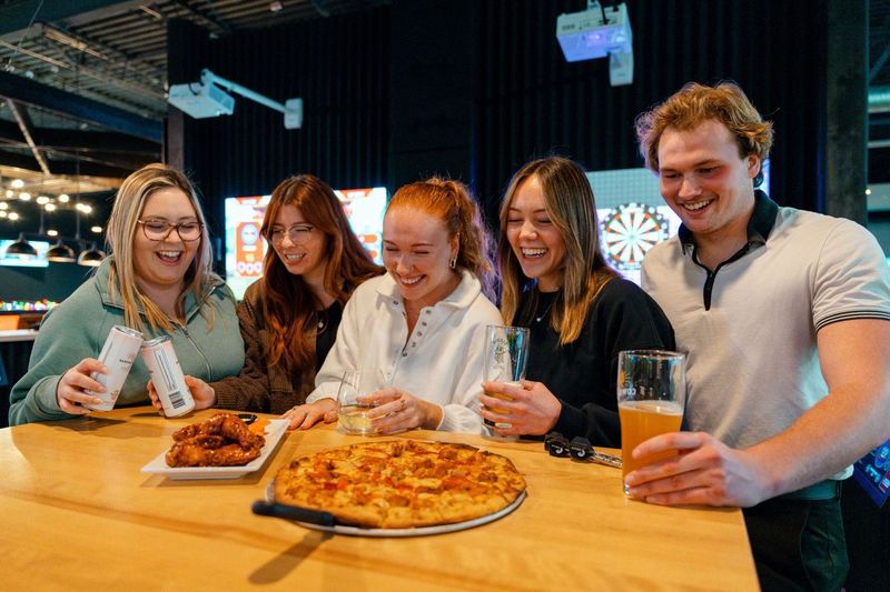Golfplay Five people stand around a table with pizza, chicken wings, and drinks, smiling and laughing in a casual setting. Dartboards and a state-of-the-art Golf Simulator from Golfplay are visible in the background. Indoor Golf