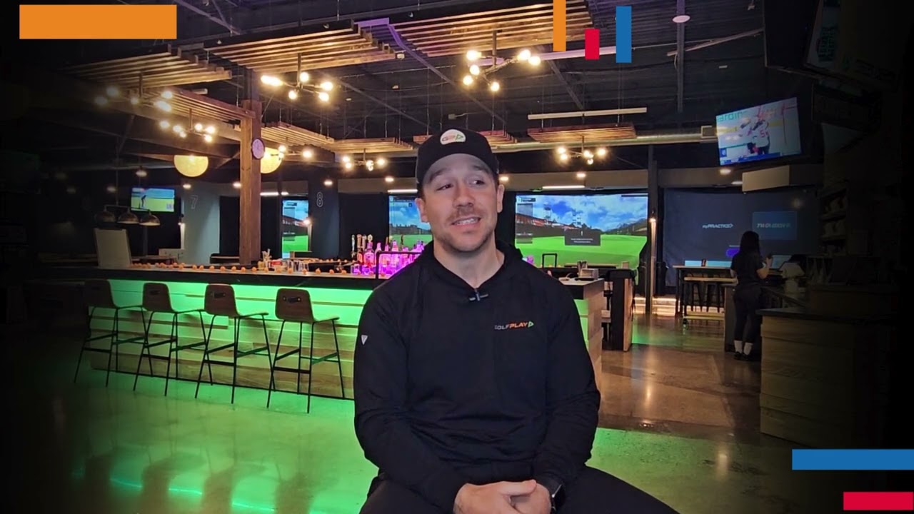 Golfplay A man wearing a black cap and shirt sits on a stool in a modern Golf Simulator venue with neon lighting and multiple screens in the background. Indoor Golf