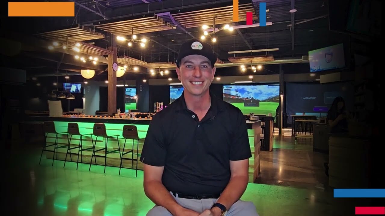 Golfplay A man in a black polo and cap sits smiling in a modern Indoor Golf entertainment venue with multiple screens, neon lighting, and a state-of-the-art Golf Simulator. Indoor Golf