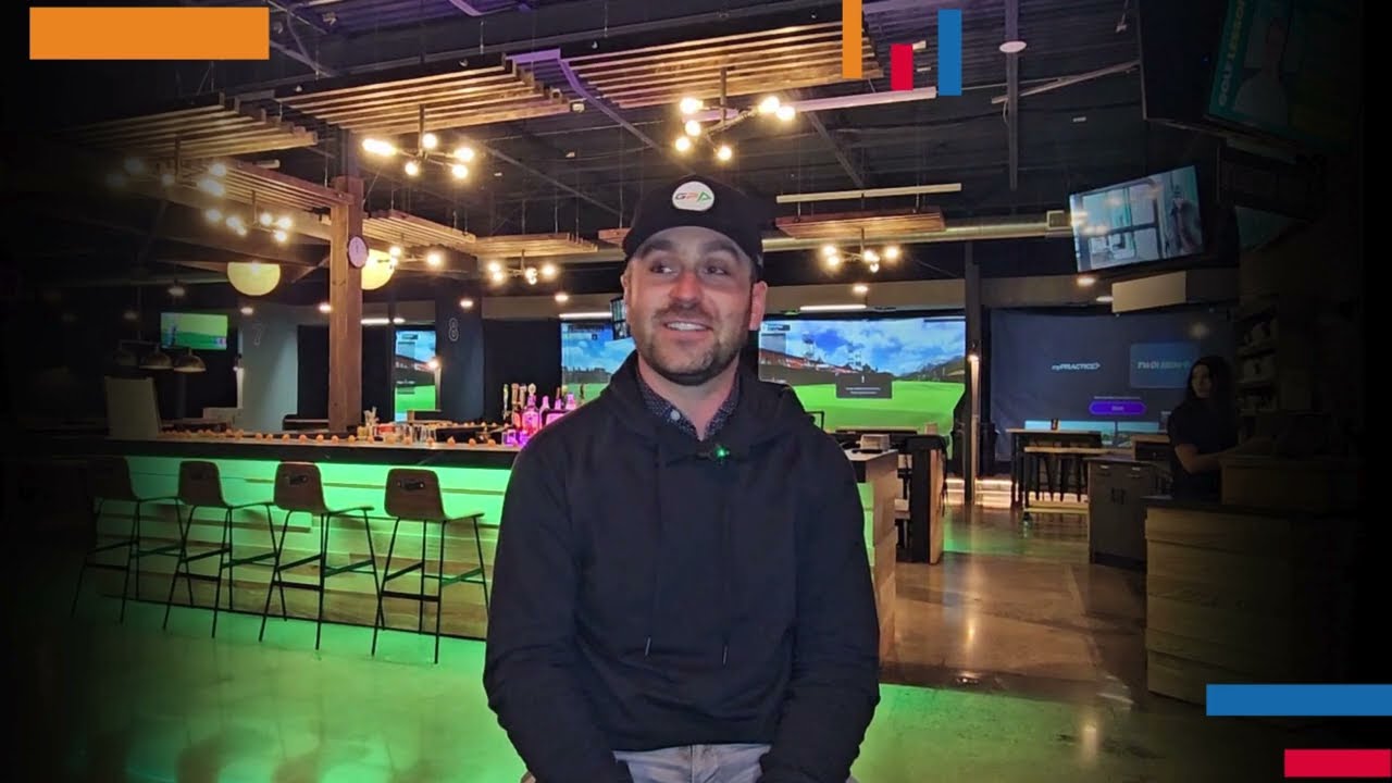 Golfplay A man wearing a cap and hoodie sits and smiles in a dimly lit, modern indoor entertainment venue with neon green lighting and overhead light fixtures, enjoying the high-tech Golf Simulator at Golfplay. Indoor Golf