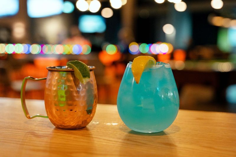 Golfplay A copper mug with a lime wedge and a round glass of blue cocktail with a lemon wedge sit on a wooden bar counter at the Golfplay indoor golf venue, with blurred lights and bar scene in the background. Indoor Golf