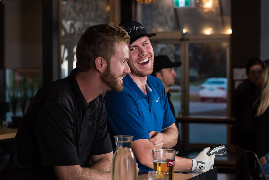 Golfplay Two men sit at a bar in Golfplay, one in a black shirt and one in blue, smiling and laughing. A golf club, a glass of beer, and a water carafe are on the table—perfect for relaxing after a round at the indoor golf simulator. Indoor Golf