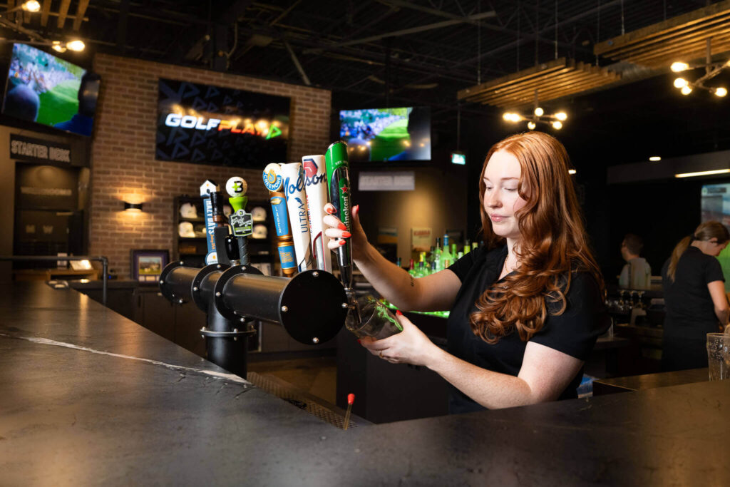 Golfplay A bartender with long red hair pours draft beer from a tap at a bar, with TV screens, patrons, and an indoor golf simulator visible in the background. Indoor Golf