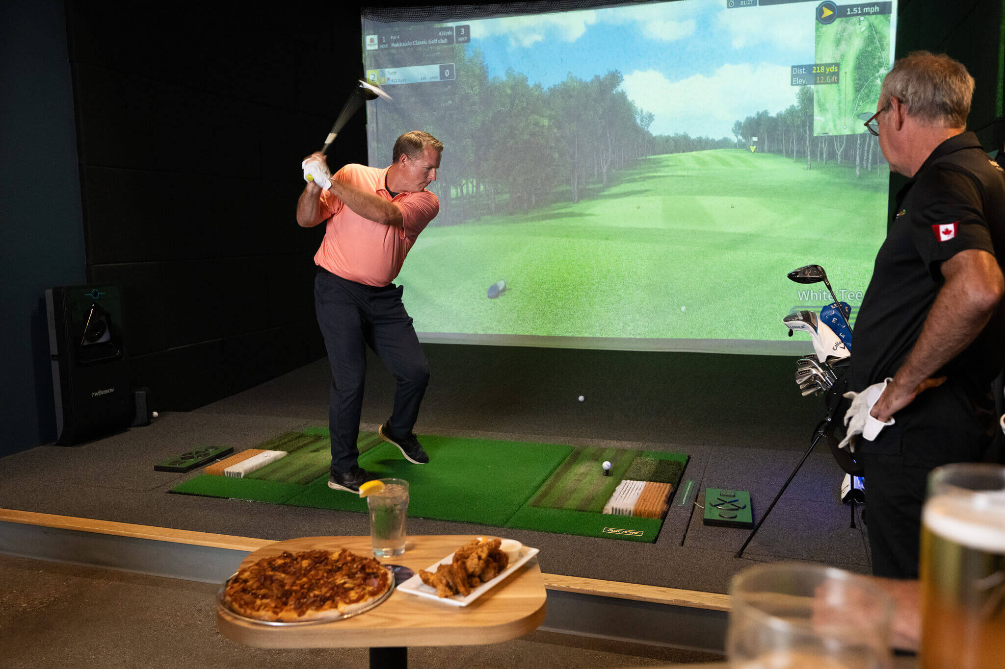 Golfplay Two men enjoy indoor golf with a Golf Simulator; one swings a club as the other watches. A table in the foreground is set with food and drinks, while a realistic golf course is displayed on the screen. Indoor Golf
