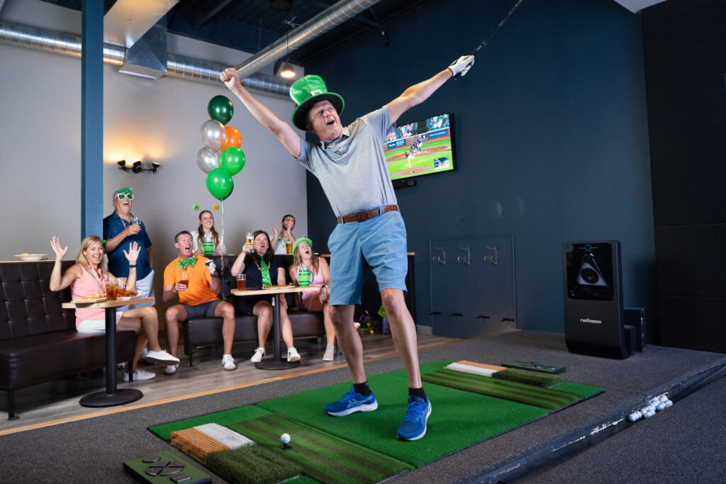 Golfplay A man wearing a green hat celebrates after swinging a golf club on an indoor golf simulator, while a group of people behind him cheer and react enthusiastically to his Golfplay. Indoor Golf
