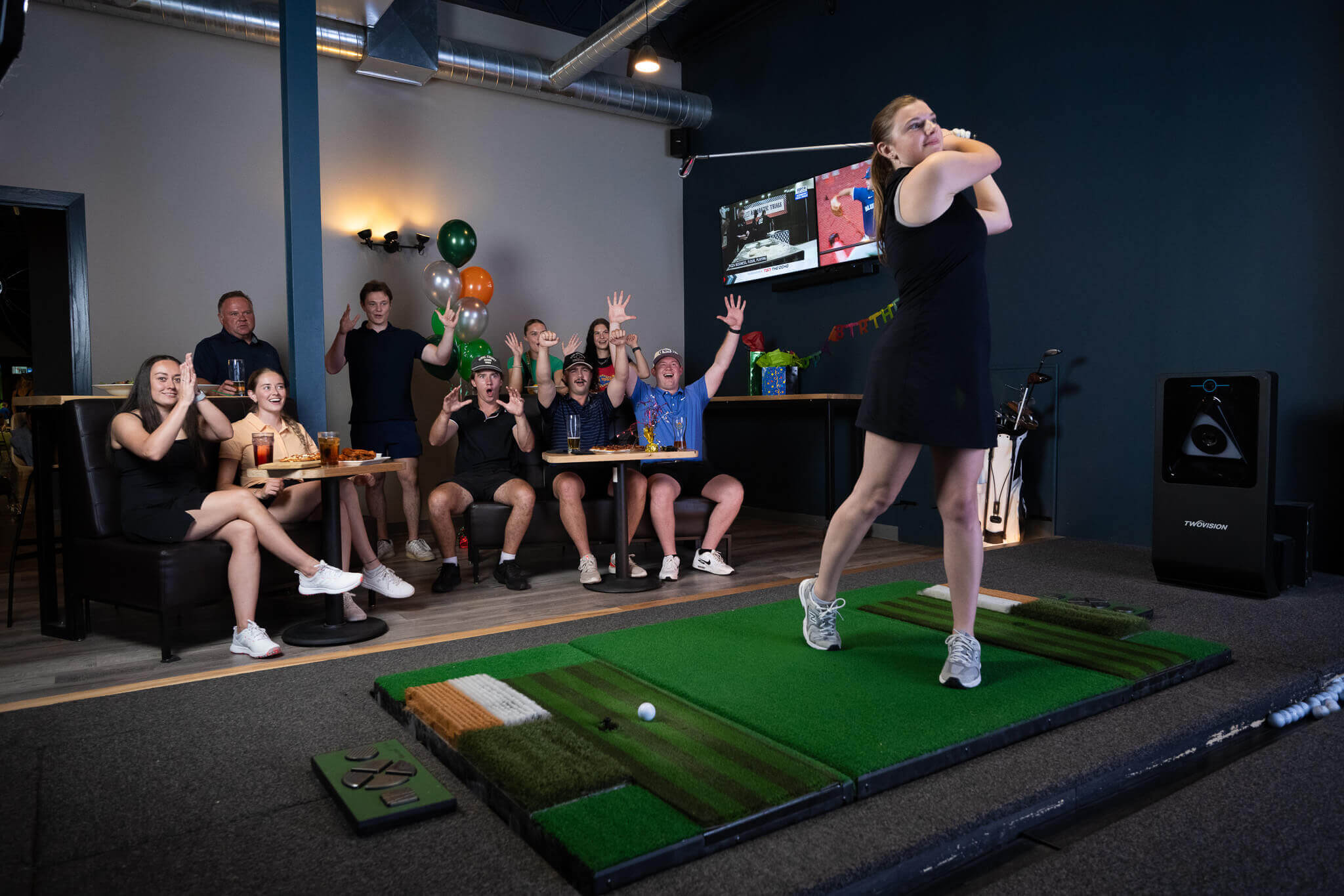 Golfplay A woman swings a golf club on a Golf Simulator as a group of people seated nearby cheer and raise their hands in excitement. Indoor Golf