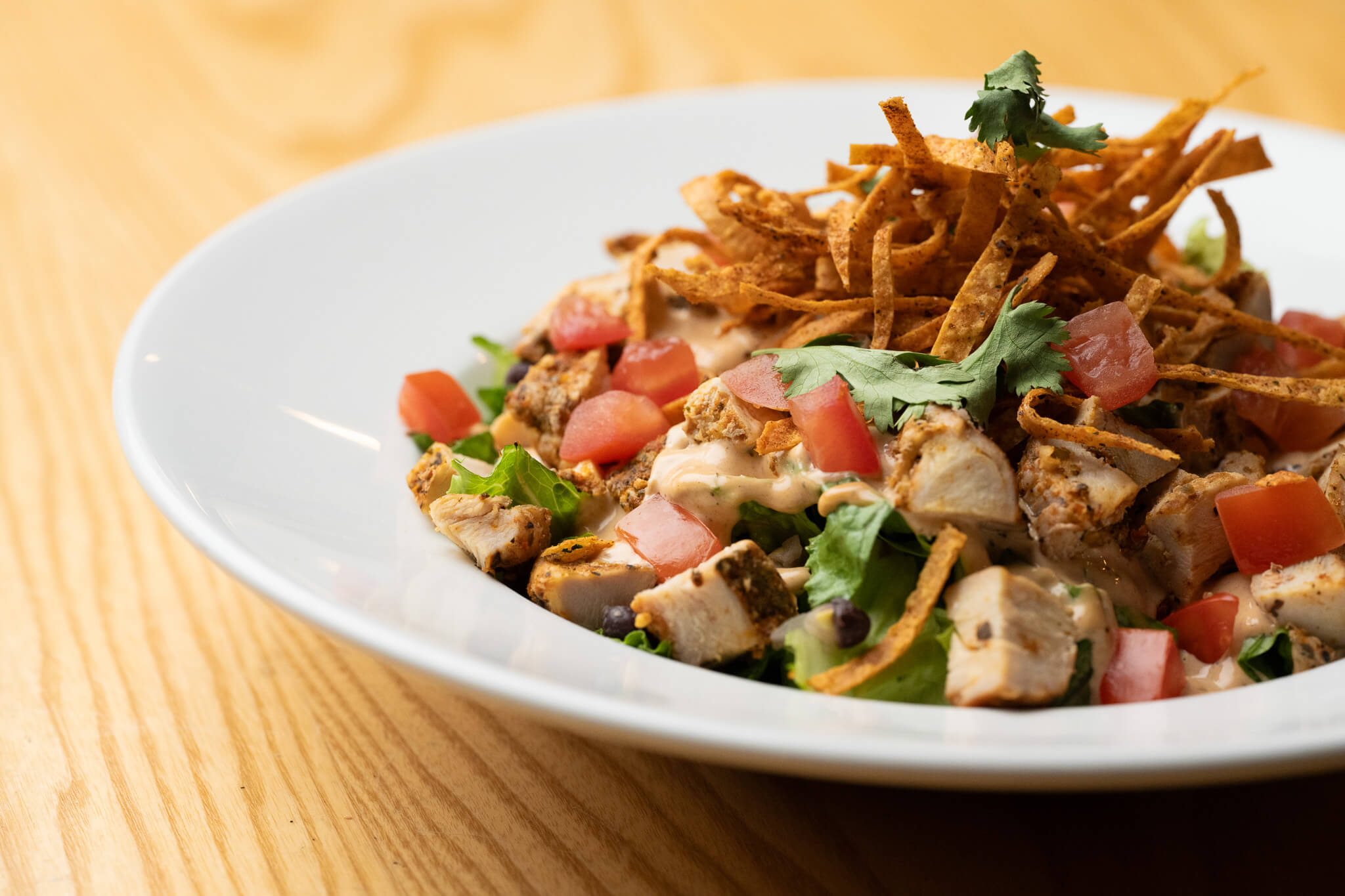 Golfplay A white plate with chopped grilled chicken, diced tomatoes, lettuce, crispy tortilla strips, cilantro, and dressing on a wooden table at Golfplay’s vibrant indoor golf simulator lounge. Indoor Golf