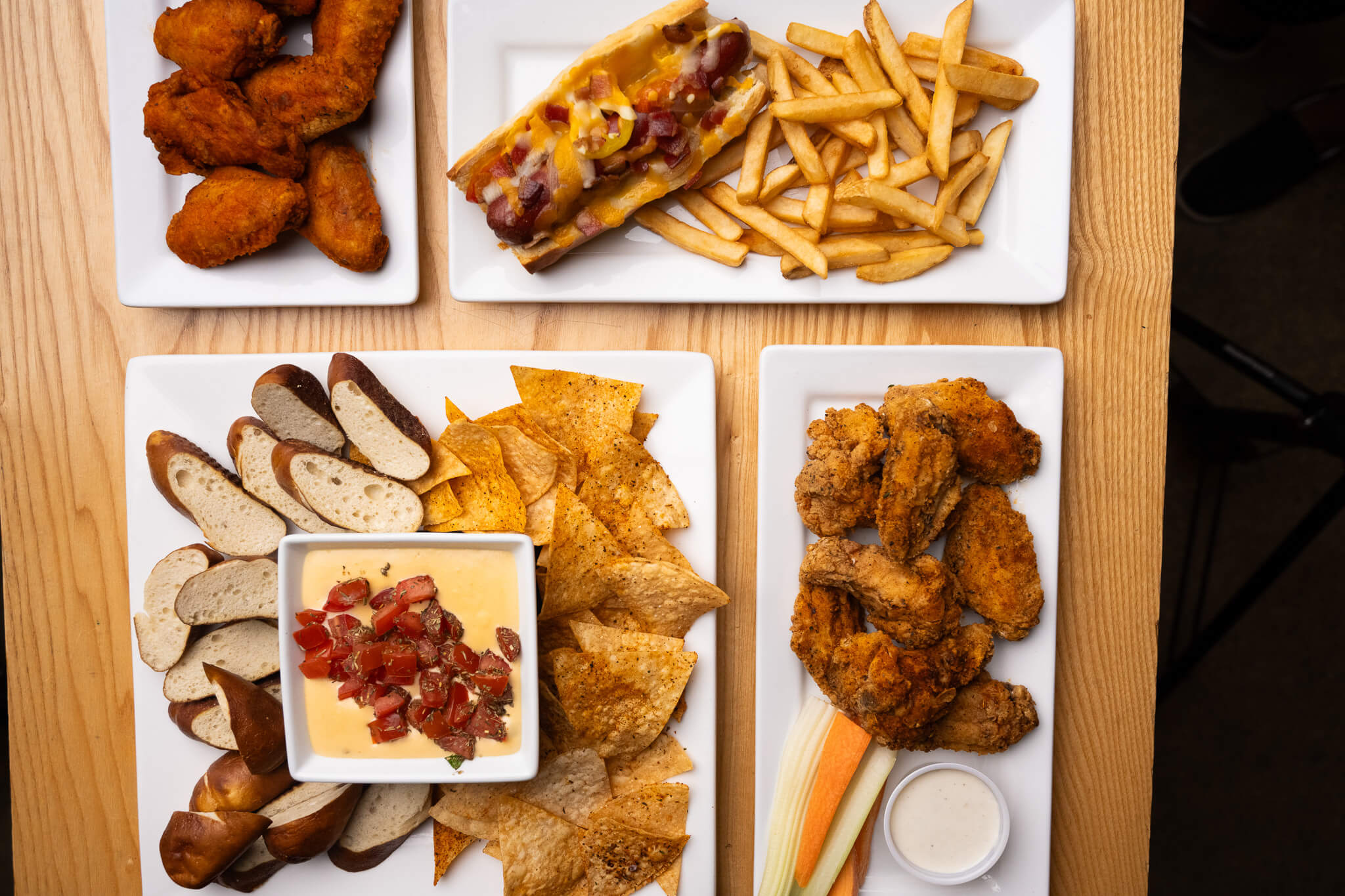 Golfplay Four plates of food on a wooden table at Golfplay: chicken wings, hot dog with fries, pretzel and chips with cheese dip, and fried chicken with celery, carrots, and ranch dip—perfect for your next Indoor Golf or Golf Simulator session. Indoor Golf