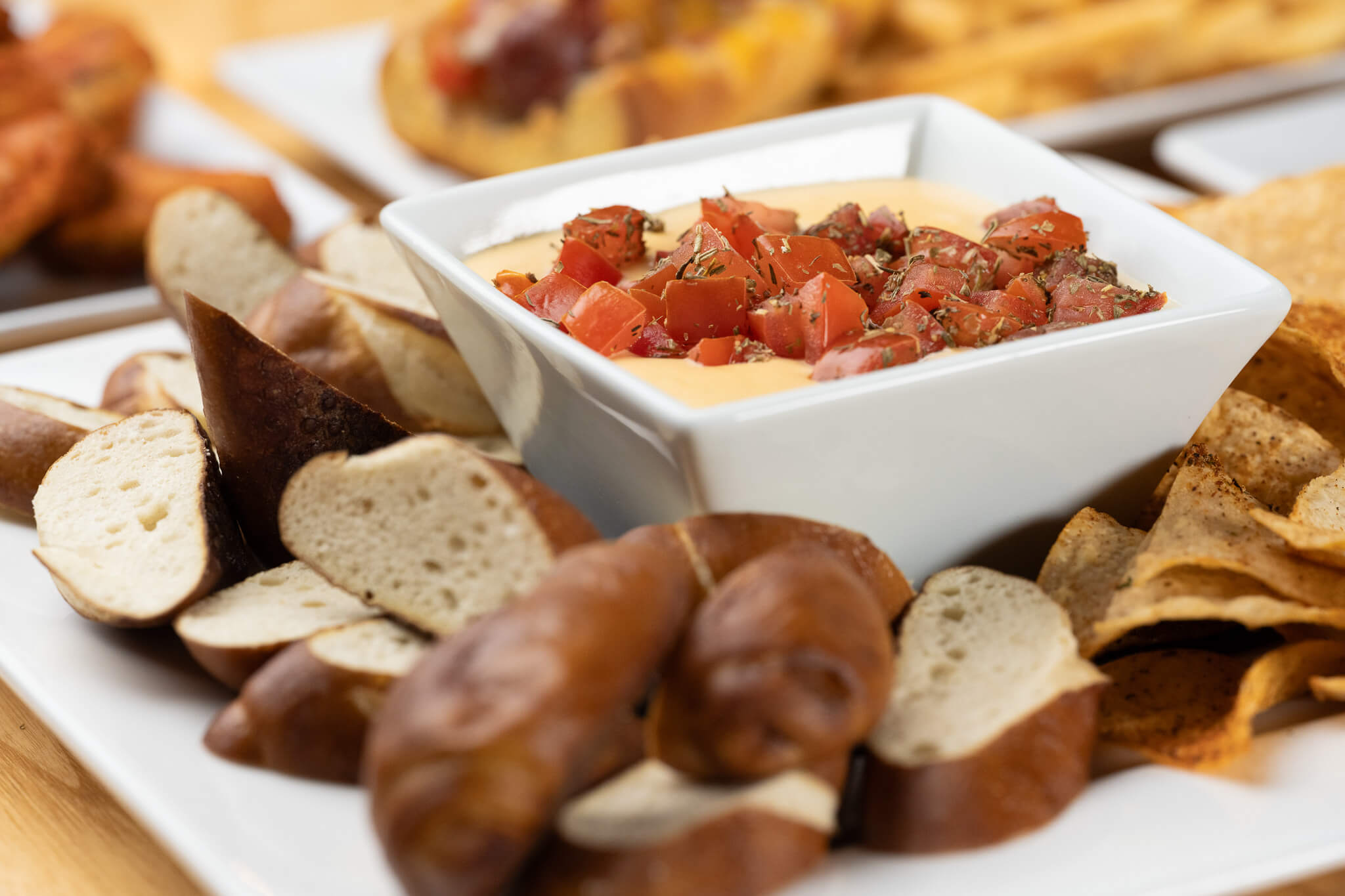 Golfplay A white square bowl of cheese dip topped with diced tomatoes and herbs, surrounded by soft pretzel pieces and tortilla chips on a white plate—perfect snack fuel for your next Indoor Golf session at Golfplay. Indoor Golf