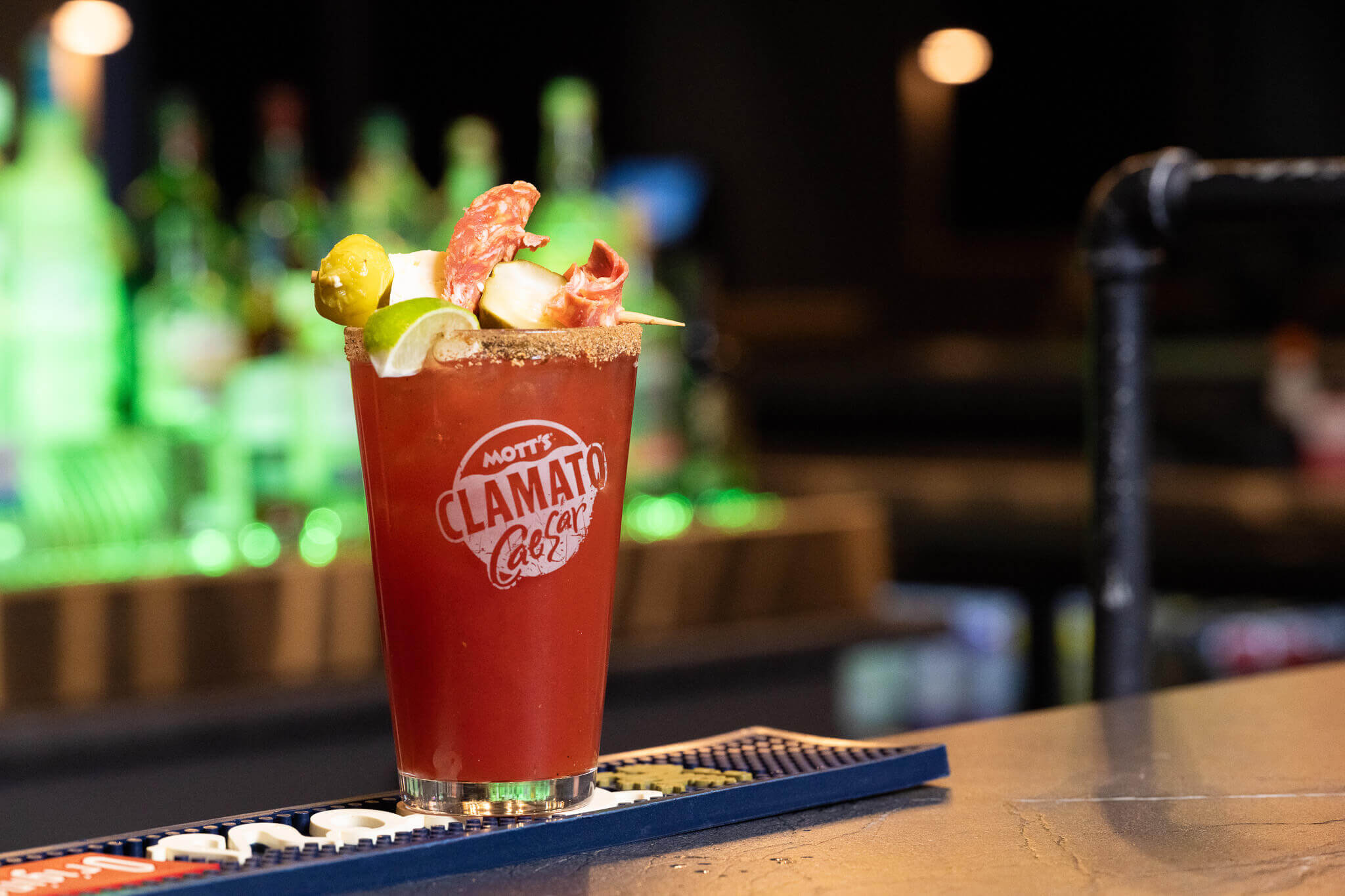 Golfplay A glass of Mott’s Clamato Caesar cocktail garnished with lime, olives, celery, and meats sits on a bar counter at an indoor golf venue, with liquor bottles blurred in the background. Indoor Golf