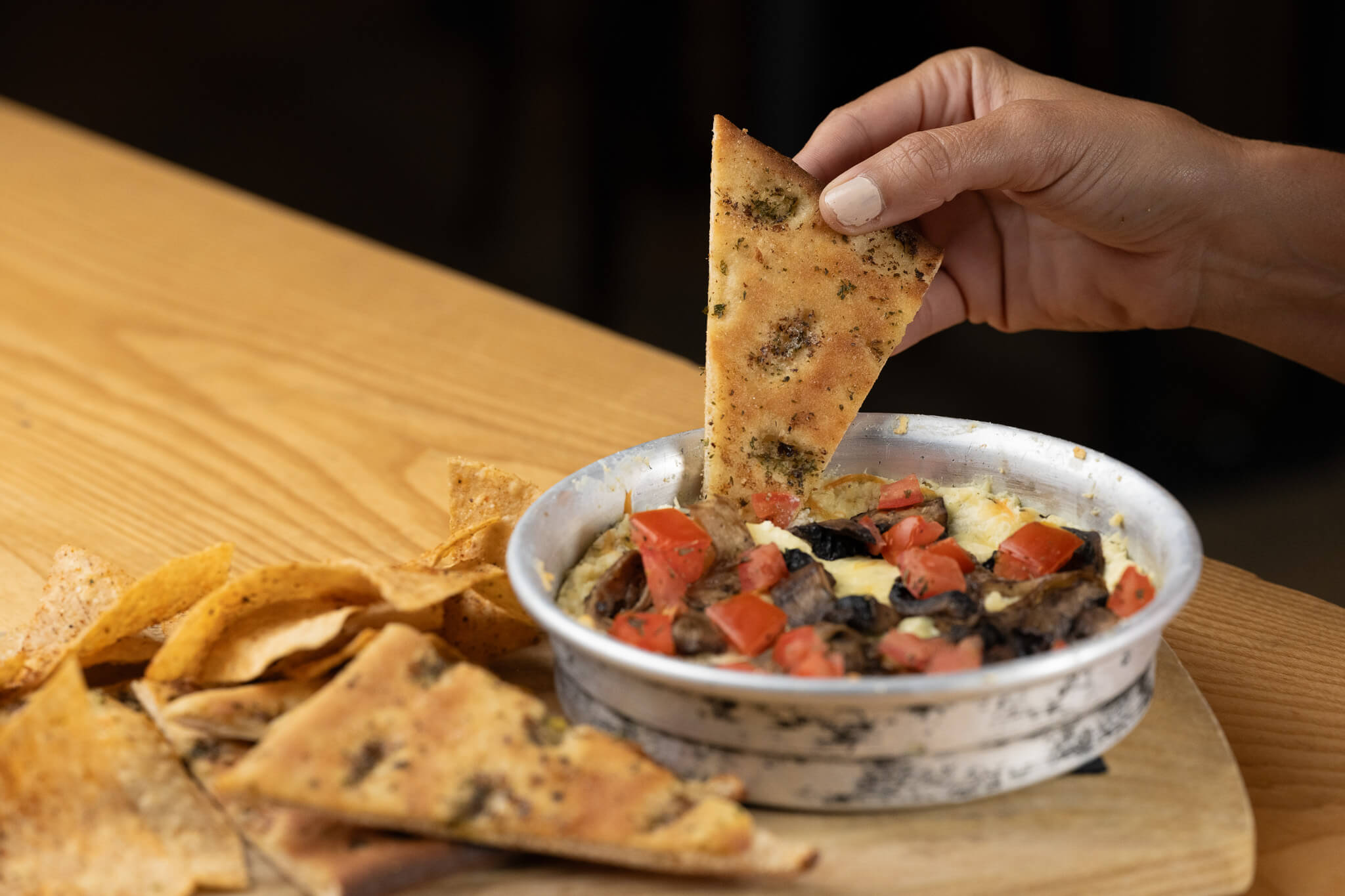 Golfplay A hand dips a piece of seasoned flatbread into a metal dish of vegetable dip, with tortilla chips and more flatbread on the side—perfect for snacking between rounds at Golfplay's state-of-the-art indoor golf simulator. Indoor Golf