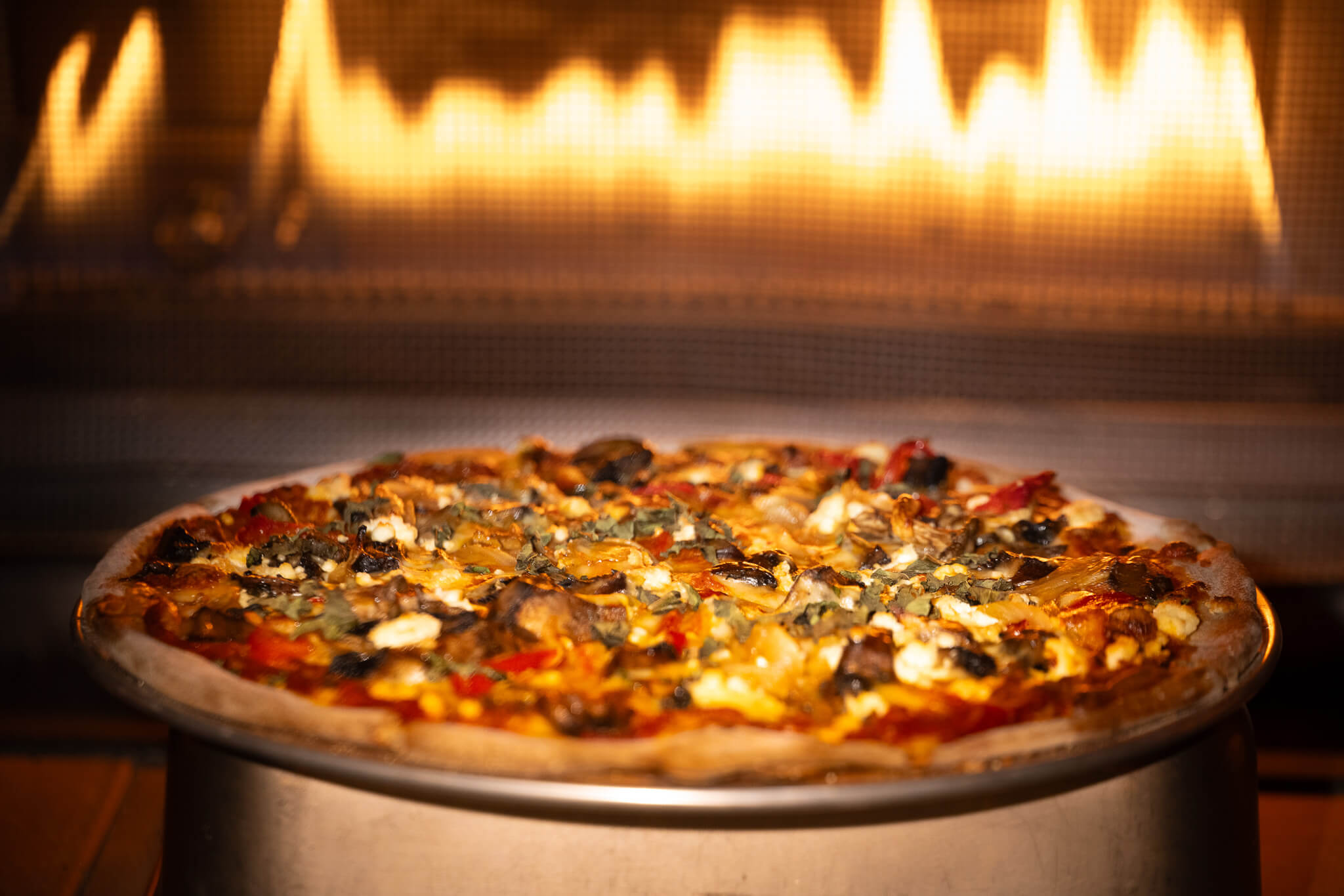 Golfplay A baked pizza topped with vegetables and cheese sits on a metal tray in front of a fireplace with visible flames, perfect for enjoying after a fun session at the nearby indoor golf simulator. Indoor Golf