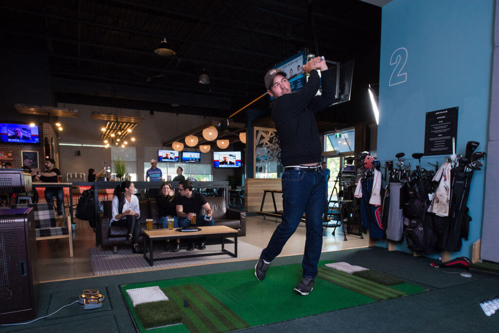 Golfplay A man swings a golf club at an Indoor Golf simulator while people sit and watch in a modern lounge setting. Golfplay equipment and golf bags are visible on the right. Indoor Golf