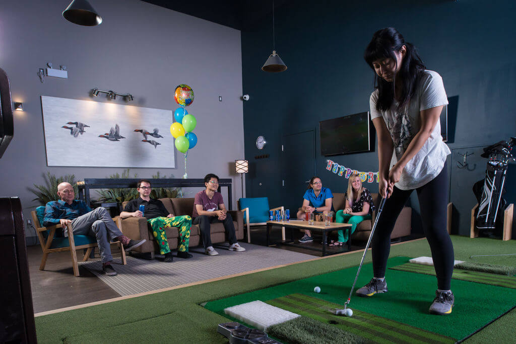 Golfplay A woman putts a golf ball on an indoor green during an Indoor Golf party while five people watch from a couch; balloons and a birthday banner are in the background. Indoor Golf